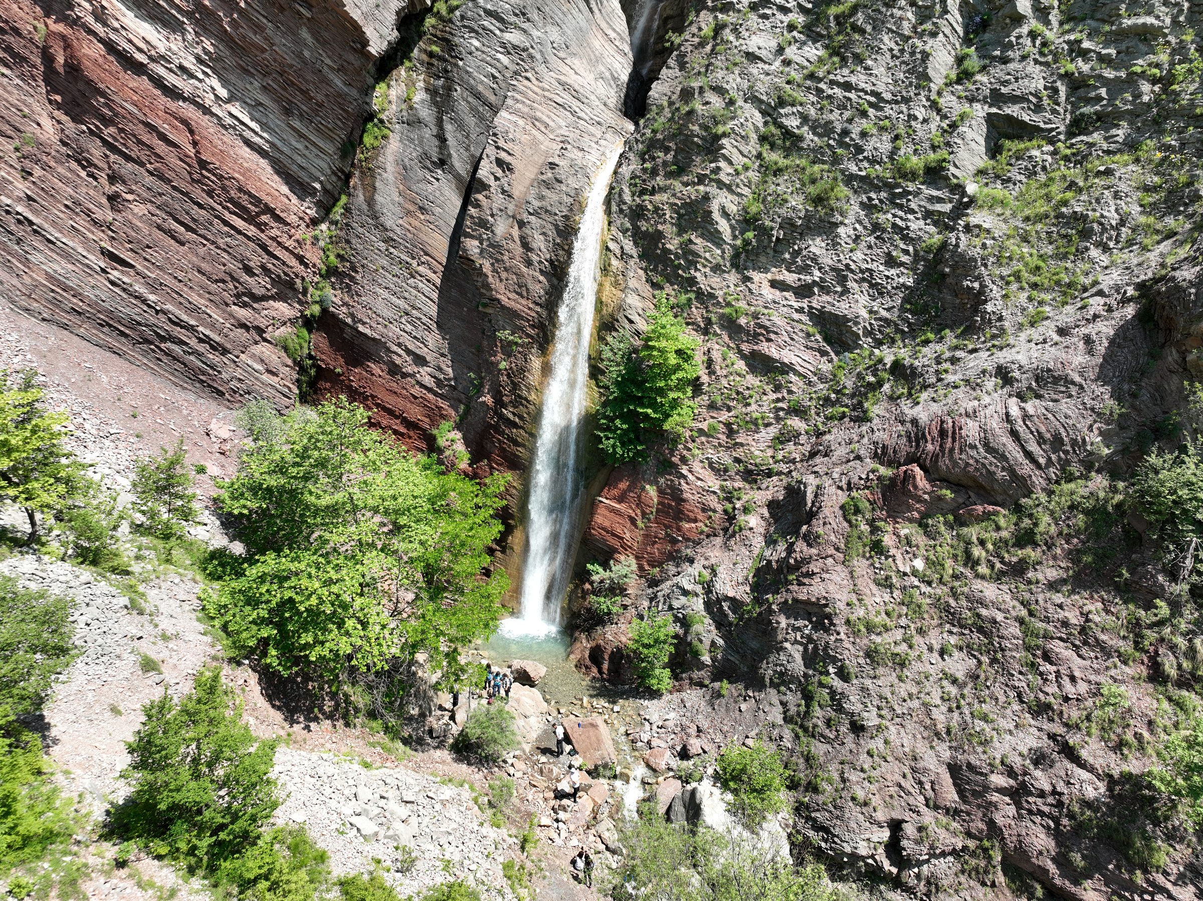 The Shëngjergj waterfall dropping through a red-rock canyon, hikers at its base