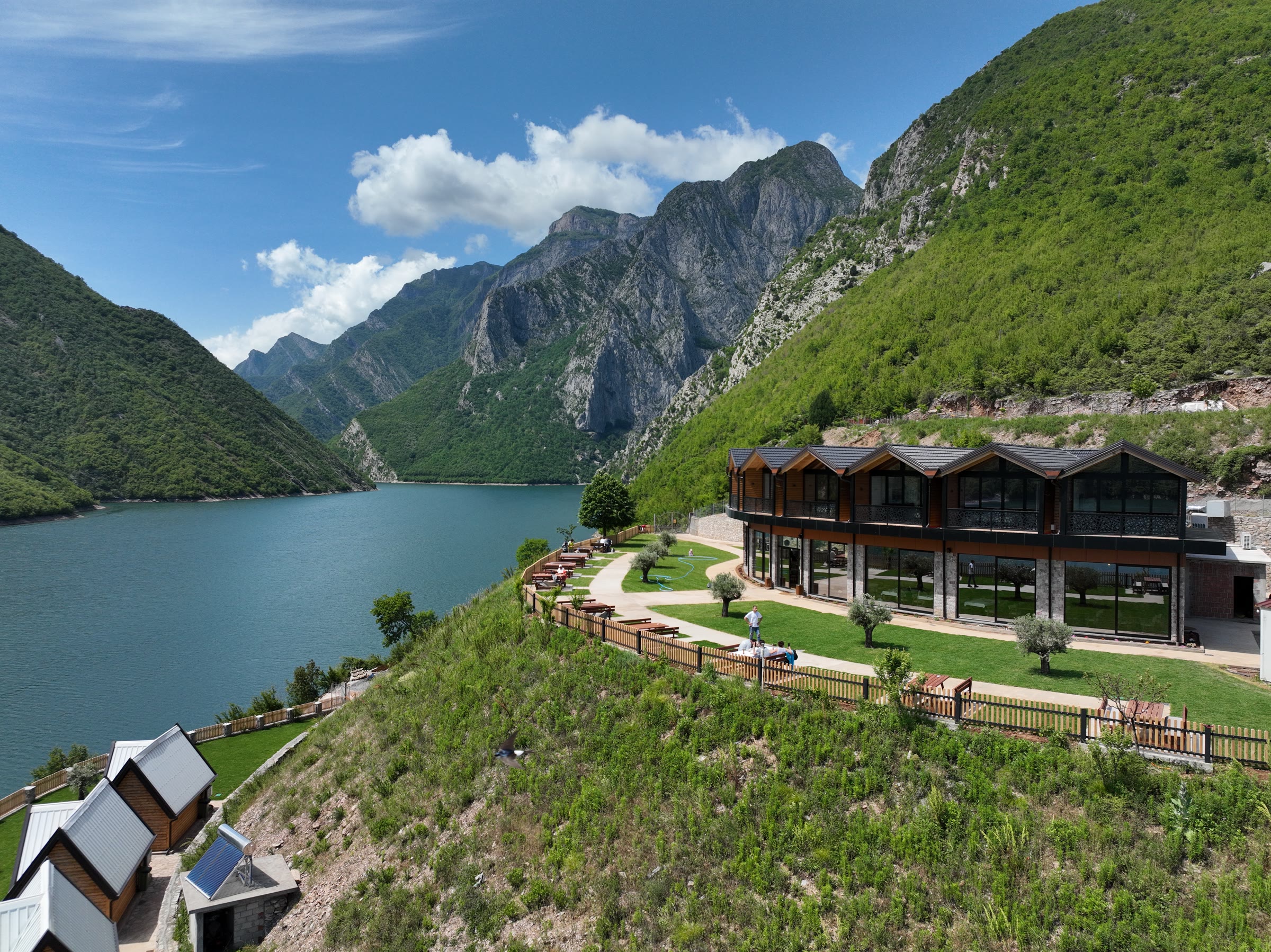 Aerial view of a lakeside guesthouse with wooden chalets, olive trees and a solar water heater, set into a mountainside above a lake