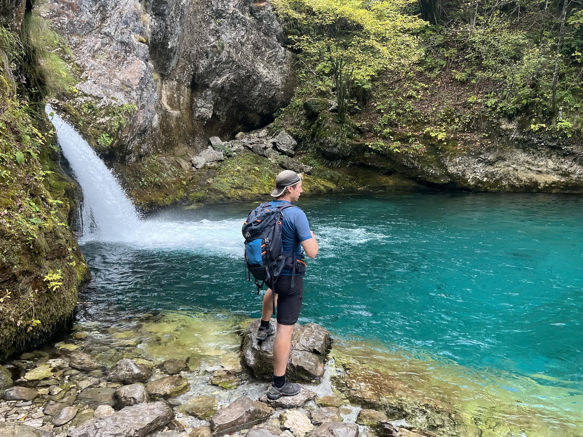 A hiker with a backpack pausing beside a turquoise pool beneath a forest waterfall in Albania